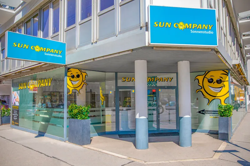 Eckgebäude mit blauen Sun Company Sonnenstudio-Schildern, Glasfassade, lachenden Sonnenmotiven und Pflanzenkübeln vor dem Eingang