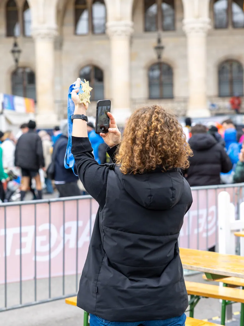 Frau mit Locken fotografiert mit dem Smartphone eine goldene Finisher-Medaille vor einer Absperrung bei einer Laufveranstaltung