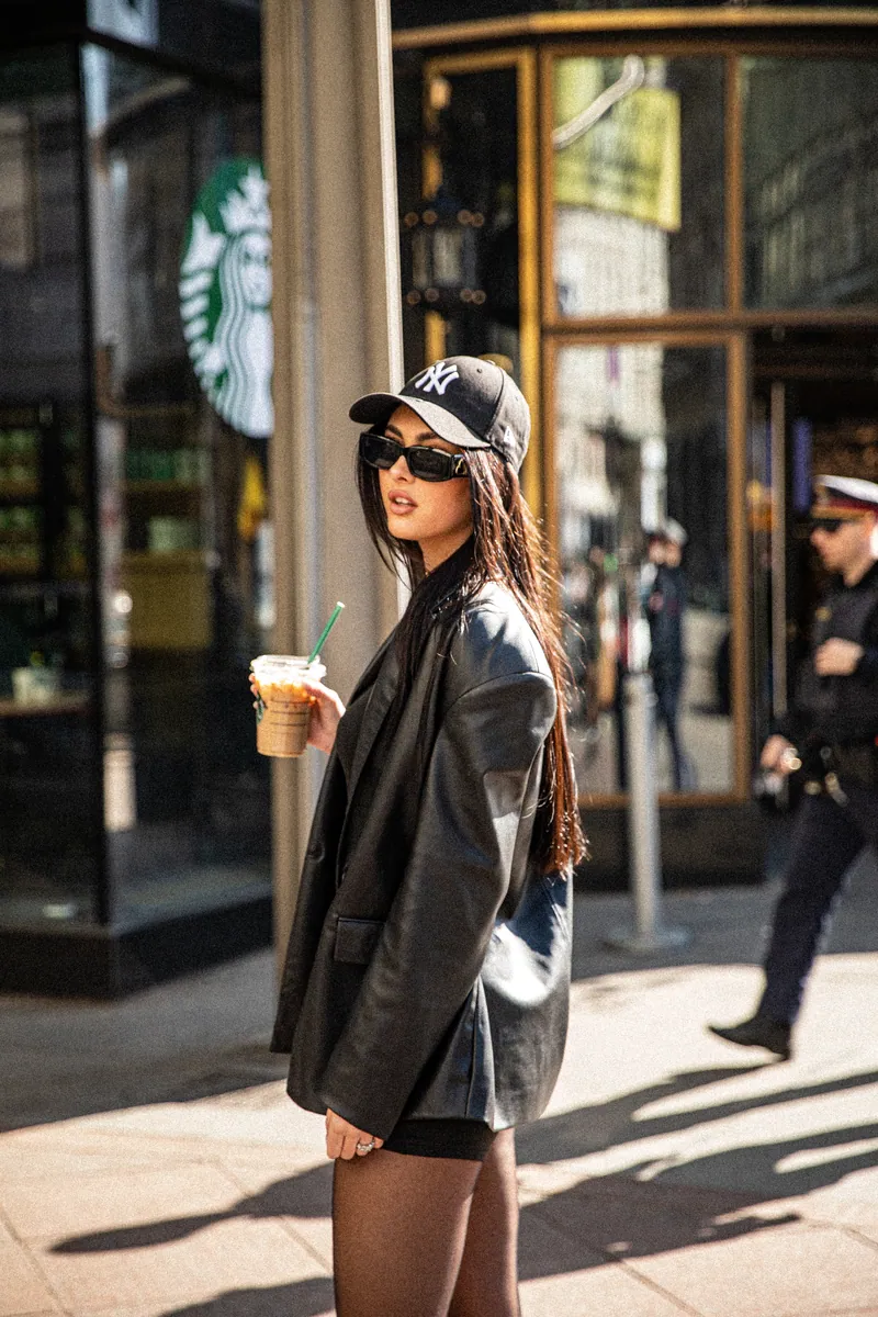 Frau mit New-York-Yankees-Kappe und Lederblazer hält einen Starbucks-Becher auf sonniger Straße