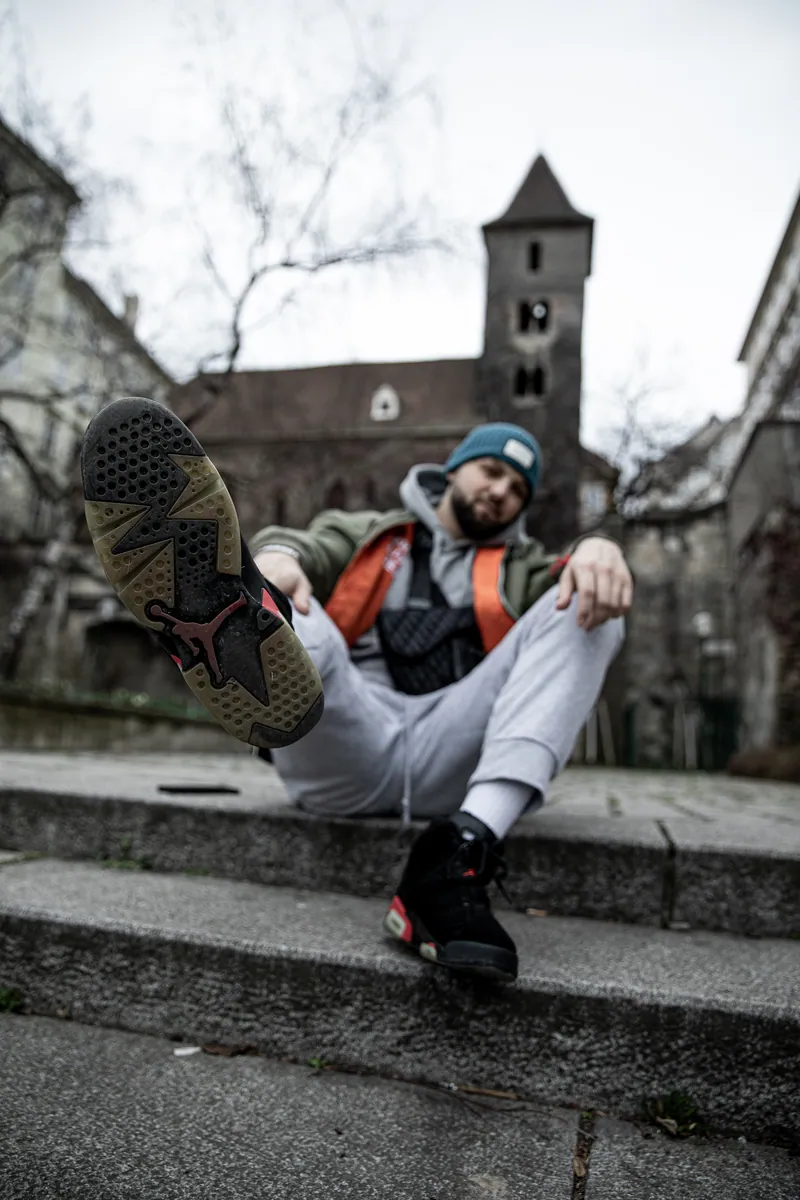 Mann mit Mütze sitzt auf Steinstufen und streckt den Jordan-Sneaker zur Kamera, dahinter ein Kirchturm