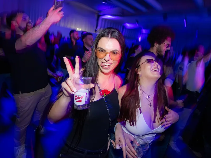 Zwei lachende Frauen feiern im Club, eine hält einen Stiegl-Becher und zeigt das Victoryzeichen